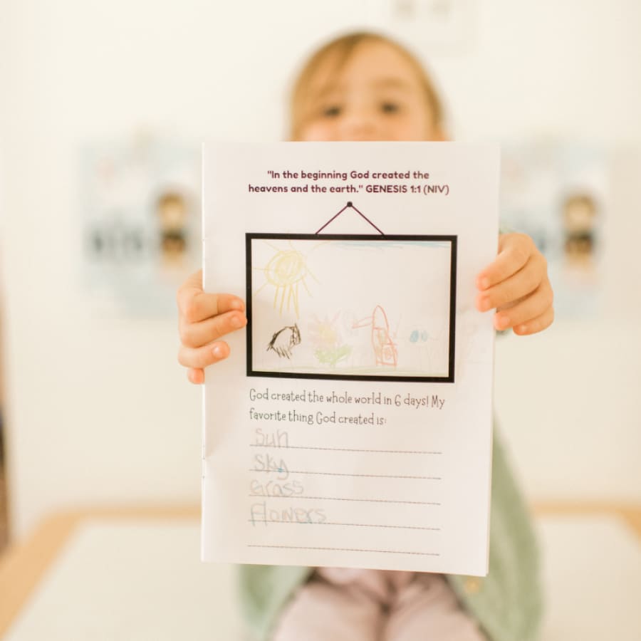 little girl showing her bible journal for kids drawing while sitting on a desk
